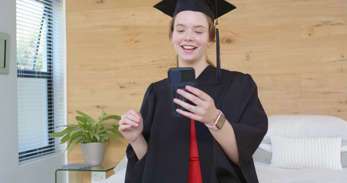 Laureata sorridente che tiene il telefono e saluta mentre si sistema il tocco, partecipando a una videochiamata di laurea dal letto.