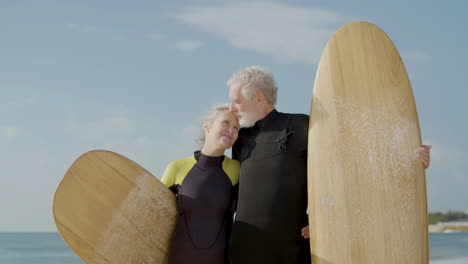 vista frontal de una pareja mayor en traje de neopreno con tabla de surf de pie en la playa de arena, mirando a la cámara y besándose