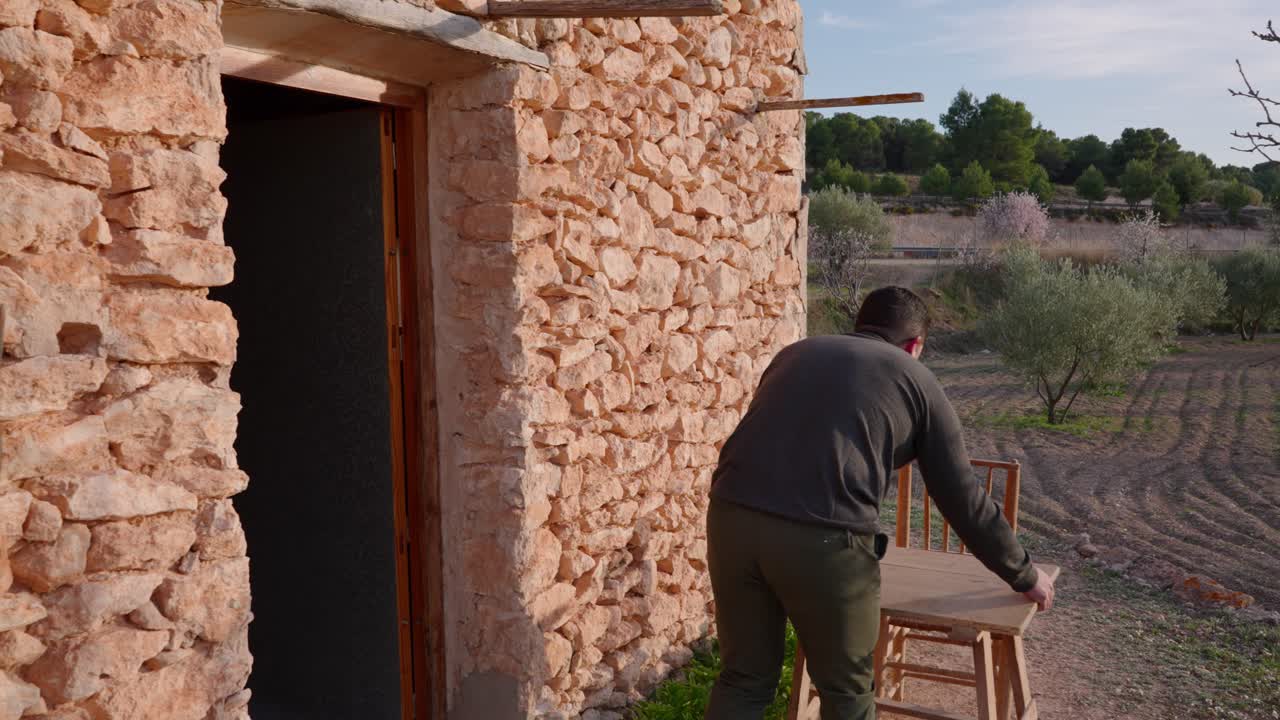 지중해 올리브 나무 사이에 있는 오래된 석조 집에서 한 남자가 시골의 이른 봄에 식탁을 준비하면서 나옵니다.