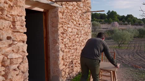 A-man-comes-out-of-an-old-stone-house-among-Mediterranean-olive-trees,-preparing-a-table-for-a-meal,-in-the-early-spring-in-the-countryside