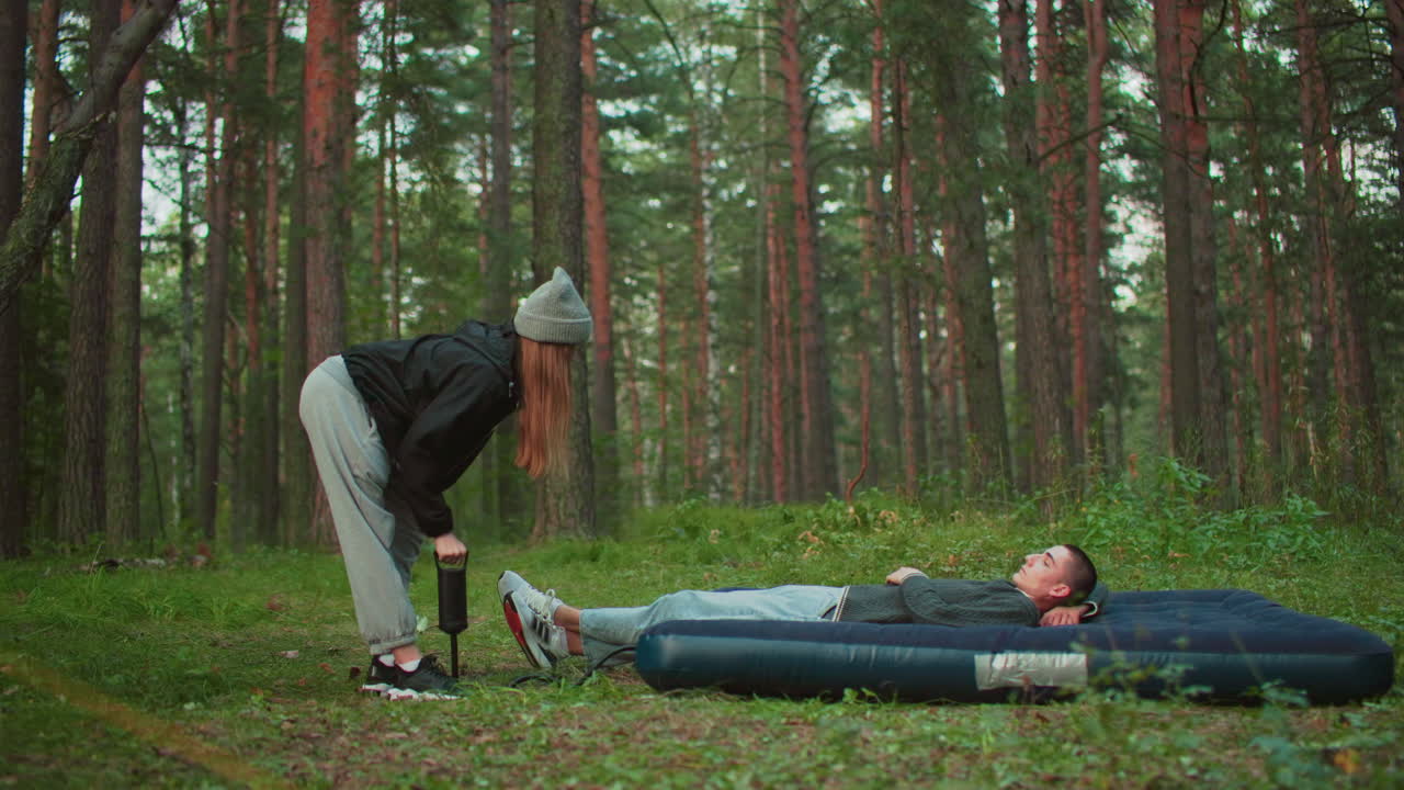 white man lies on air bed while woman pumps air into it with pitched tent behind in forest clearing surrounded by trees and soft daylight