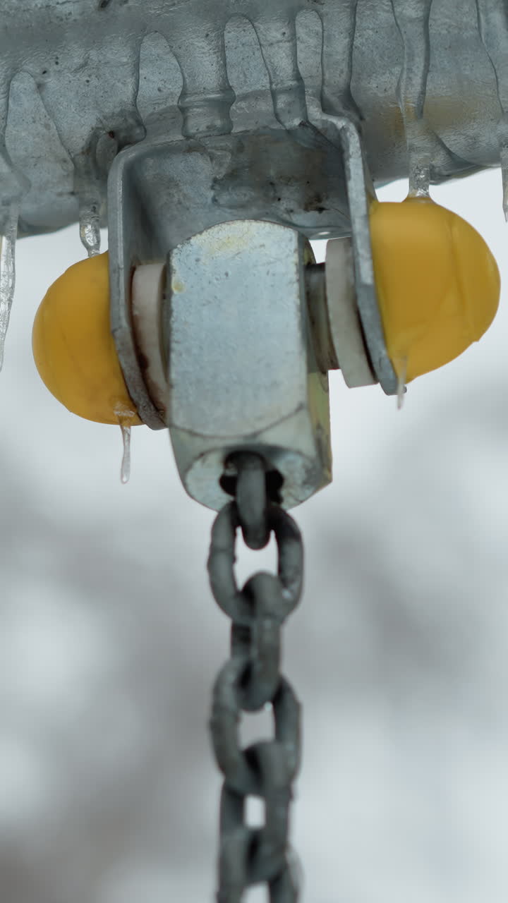 primer plano de la articulación metálica de la cadena oscilante con tapón amarillo recubierto de hielo y hielos colgando a lo largo de una barra de metal, destacando texturas congeladas y detalles invernales contra un fondo nevado borroso