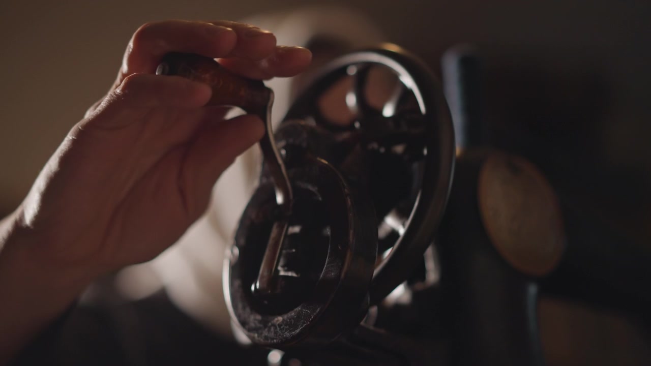Mulher concentrada ajustando delicadamente uma máquina de costura histórica sob uma luz quente.
