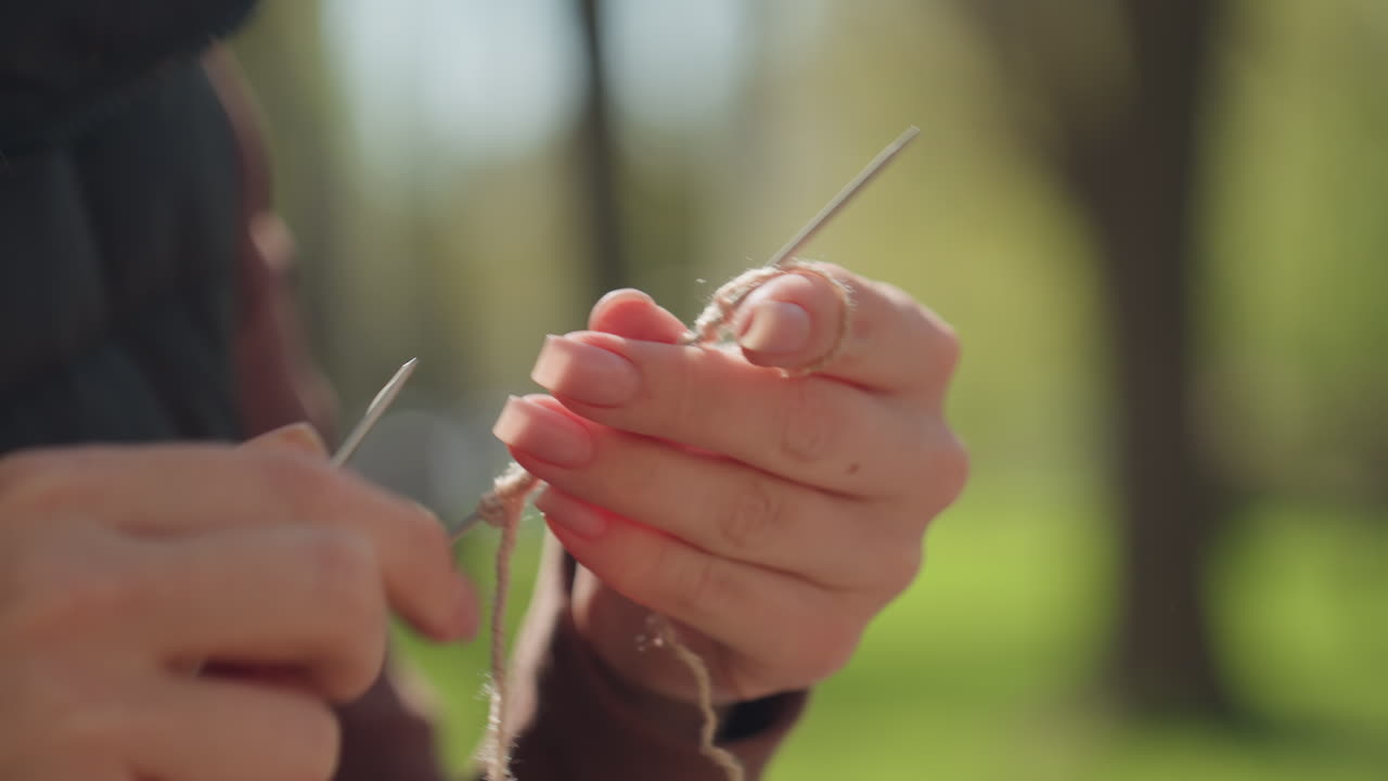 아늑한 니트 스카프를 만드는 여성의 클로즈업, 잎이 무성한 나무 아래에서 부드러운 스카프를 꼼꼼하게 뜨개질하는 평화로운 순간, 여성이 실외에서 양모 액세서리를 손으로 만드는 고요한 장면