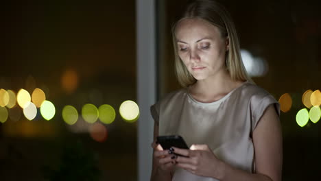 vista de cerca de una mujer rubia escribiendo un mensaje en un smartphone en la oscuridad con luces desenfocadas en el fondo