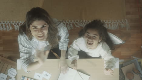 top down of jolly girl and mother studying at home and waving at camera