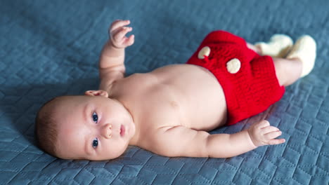 baby boy lying on face up in red pants with white buttons and socks. lovely infant kid staying calm and peaceful. close up.