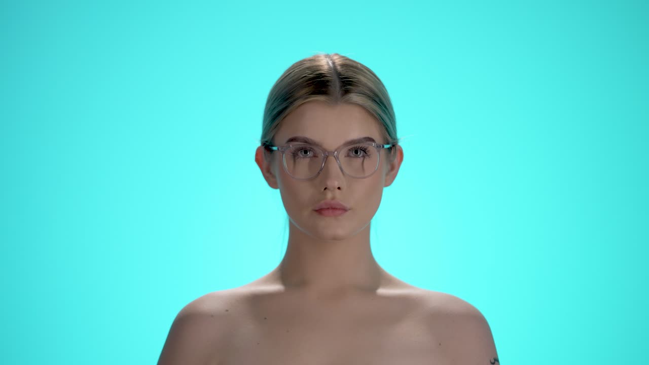 Medium static shot of a young pretty beautiful woman putting on her glasses for a clear view and looking at the camera in front of turquoise background in slow motion