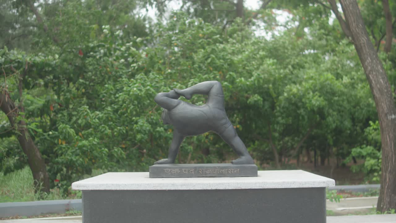 Slow pull-out shot features a dark stone statue of Eka Pada Rajakapotasana (One-Legged King Pigeon Pose) on a pedestal inscribed in Hindi, surrounded by vibrant greenery in a tranquil Indian garden
