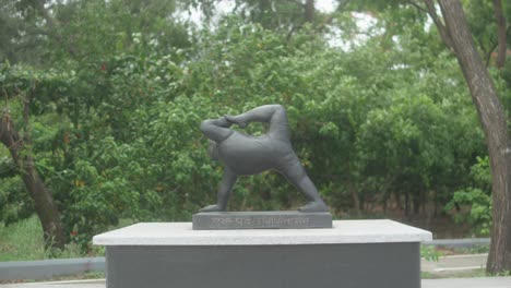 slow pull-out shot features a dark stone statue of eka pada rajakapotasana (one-legged king pigeon pose) on a pedestal inscribed in hindi, surrounded by vibrant greenery in a tranquil indian garden