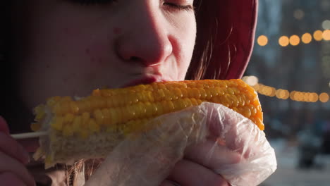 close-up of young girl eating corn held by stick wrapped in white plastic with bokeh lights in blurred background, steam rising from fresh corn in outdoor setting