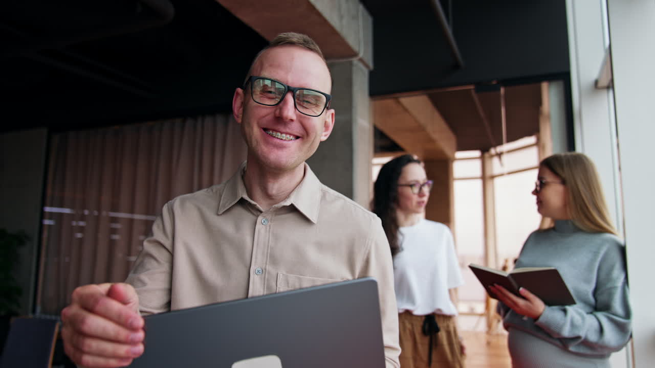 Happy cheerful man in glasses finishes work on his laptop and closes it. Female colleagues at backdrop talk standing at the window.