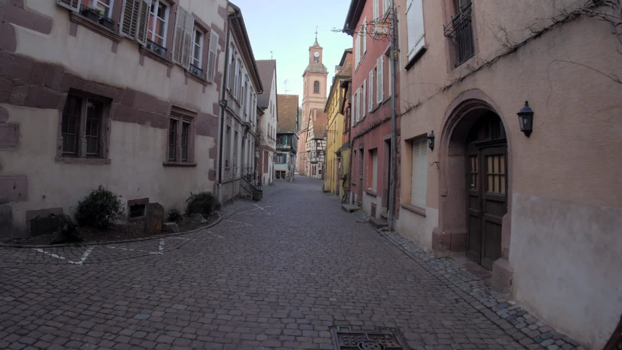 Filmato cinematografico del borgo medievale di Riquewihr, in Francia. La telecamera segue una strada acciottolata e tortuosa, superando facciate storiche colorate, verso un campanile in lontananza