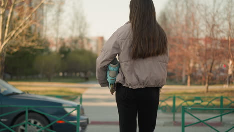 close-up back shot of a girl wearing a peach jacket and black pants, holding a turquoise skateboard, standing on a park pathway in early spring with bare trees and a blurred urban background