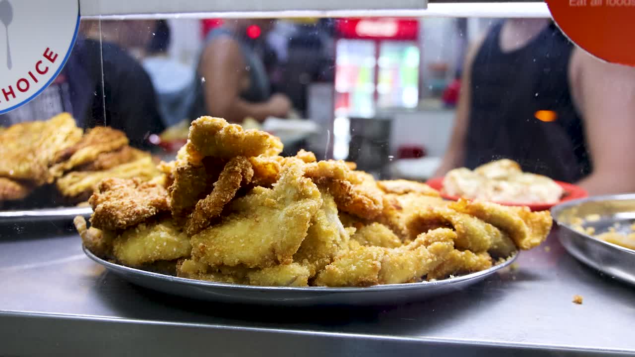 A vendor serves deep fried pork cutlets at a brightly lit Singapore hawker center, with close-up focus on the plated food and bustling background