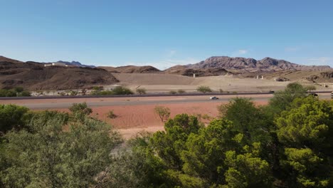 Side-View-of-Highway-in-Desert-with-Trees-in-Foreground,-Blue-Skies-and-Mountains-in-Background