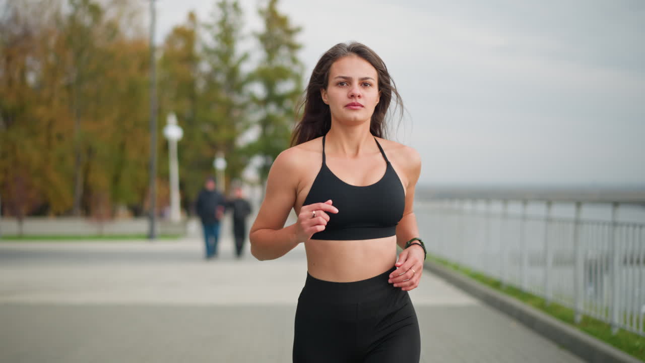 Primo piano di una giovane donna in abbigliamento fitness nero che fa jogging in un parco con sfondo sfocato che mostra un uomo che abbraccia la moglie, alberi, una recinzione di ferro e un fiume, che trasmette energia fitness all'aperto.