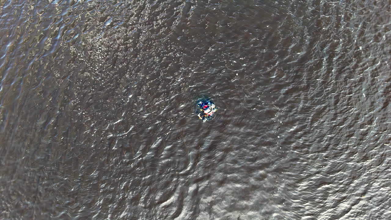 Diver in wetsuit and full scuba gear with red and blue accents floats calmly on the rippling water surface, surrounded by textured waves reflecting shifting daylight