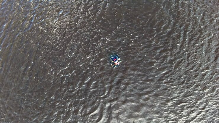 Diver in wetsuit and full scuba gear with red and blue accents floats calmly on the rippling water surface, surrounded by textured waves reflecting shifting daylight