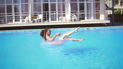 jeune fille sexy avec des lunettes de soleil, un cocktail et des dreads sur l'anneau en caoutchouc gonflable posant dans la piscine