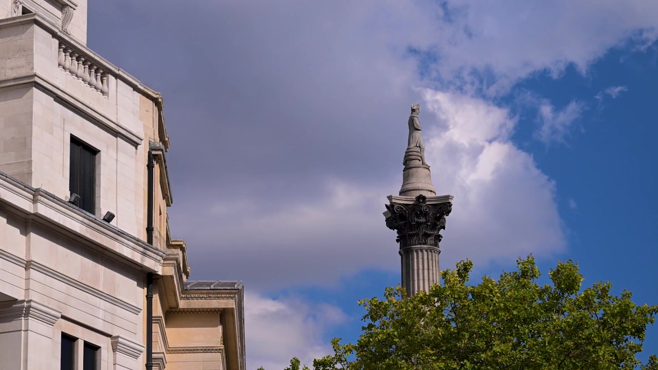 La Colonna di Nelson si erge dietro alberi frondosi accanto a un edificio in pietra chiara sotto un cielo luminoso e nuvoloso a Trafalgar Square