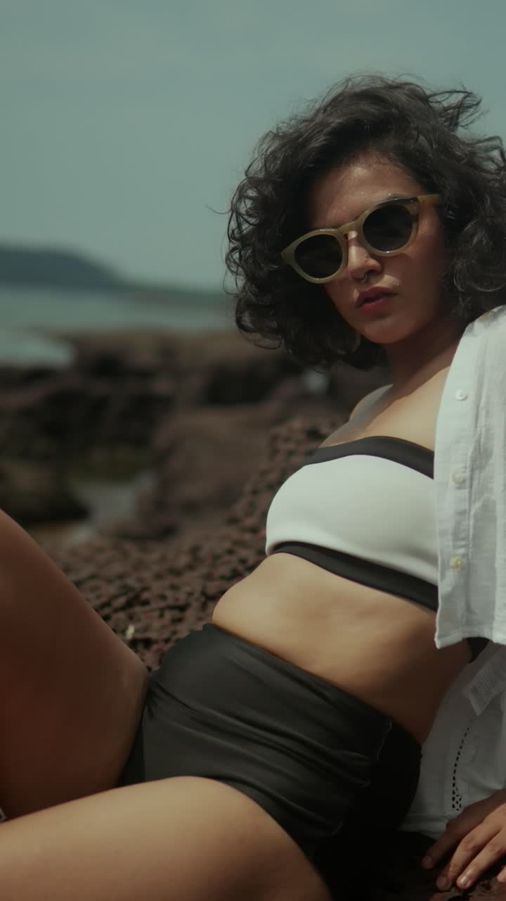 Mid-close shot of woman in sunglasses and layered beachwear posing against rocky coastline