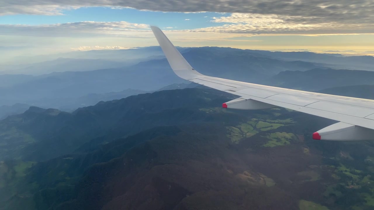 Utsikt fra flyvindu over en vinge over lagdelte fjellrygger, med varmt sollys langs horisonten under en skyet himmel