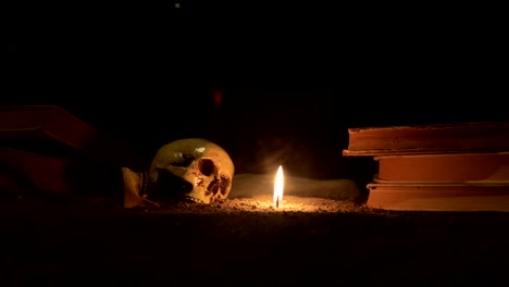 wizard's desk. a desk lit by candle light. a human skull, old books on sand surface. halloween still-life background with a different elements on dark toned foggy background. slider shot