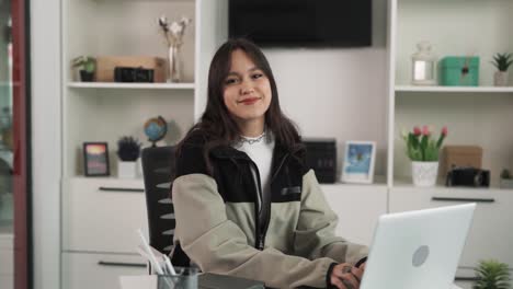 The-portrait-of-a-young-brunette-woman-who-is-focusedly-working-on-her-laptop,-smoothly-shifts-her-gaze-towards-the-camera,-and-smiles-while-looking-into-it