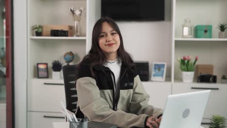 el retrato de una joven mujer morena que está enfocada trabajando en su computadora portátil, cambia suavemente su mirada hacia la cámara, y sonríe mientras la mira