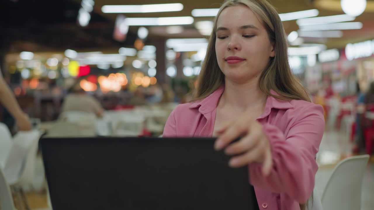 mujer en vestido rosa se sienta frente a su portátil, mirando feliz mientras lo cierra, una mujer camina en el fondo, en un restaurante bien iluminado, con una vista borrosa de la gente en el fondo