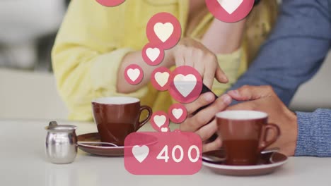 Animación-De-Iconos-De-Corazón-Flotando-Sobre-Una-Pareja-Caucásica-Usando-Un-Teléfono-Inteligente-Y-Tomando-Café