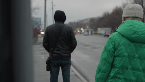 close-up of two men standing by the roadside, one wearing a green sweater and head warmer, and the other in a black sweater, with car passing by