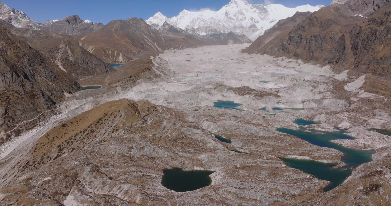 Ngozumpa Buzulu, Everest, Nepal'den panoramik bir drone yörünge çekimi; dik dağ yamaçları ve yanal moren sırtları üzerinden geçerek Cho Oyu'nun altındaki geniş buzul yatağını ve buzul üstü eriyik su göllerini gözler önüne seriyor.