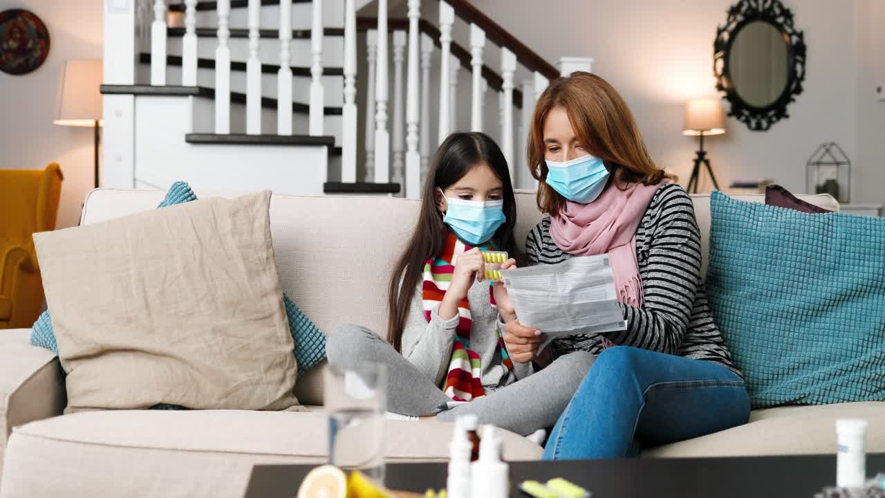 외과용 마스크를 쓰고 어머니와 함께 집에 앉아 있는 아픈 어린 소녀