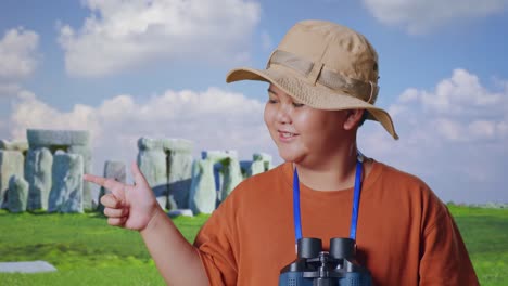 asian boy with a hat and binoculars smiling and pointing to side while traveling in stonehenge. boy researcher examines something, travel tourism adventure concept, close up
