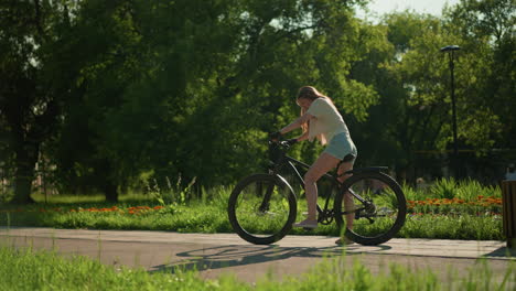 female cyclist mounts bicycle near vibrant garden, preparing to ride under warm sunlight, background displays lush greenery, colorful flower beds, and two people strolling along the garden path