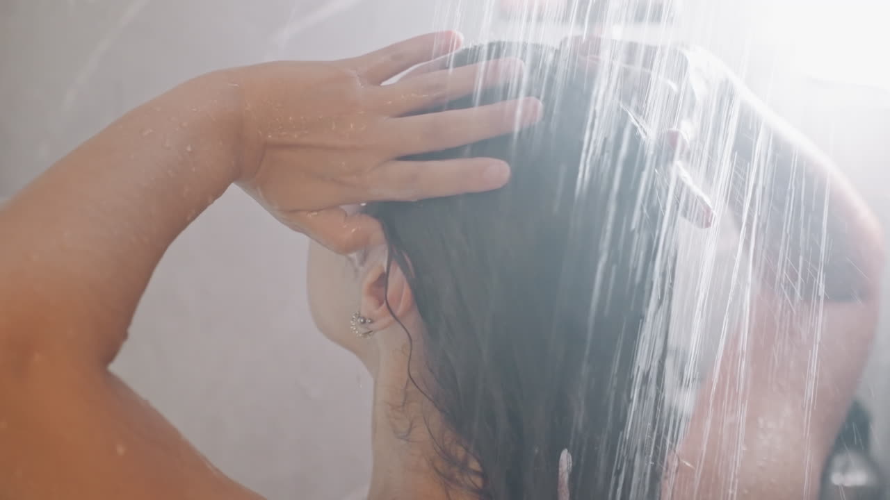 Ospite che si lava i capelli sotto la doccia. Scena ben illuminata di un'ospite che lava energicamente i capelli sotto il getto d'acqua. Azione energica di una donna che risciacqua lo shampoo dai capelli sotto la doccia con illuminazione brillante.