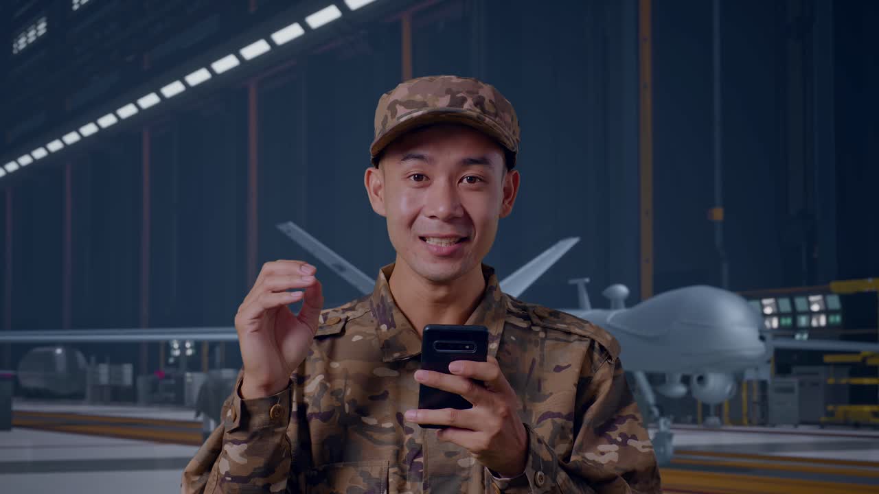 Primo piano di un soldato asiatico che guarda lo smartphone, sorride e dice "Wow" mentre è in piedi con un moderno drone militare in un hangar di manutenzione