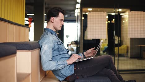 Footage-of-a-young-attractive-guy-sitting-in-an-empty-open-co-working-area,-indoors,-with-laptop-on-his-laps,-typing-something,-surfing-the-web.