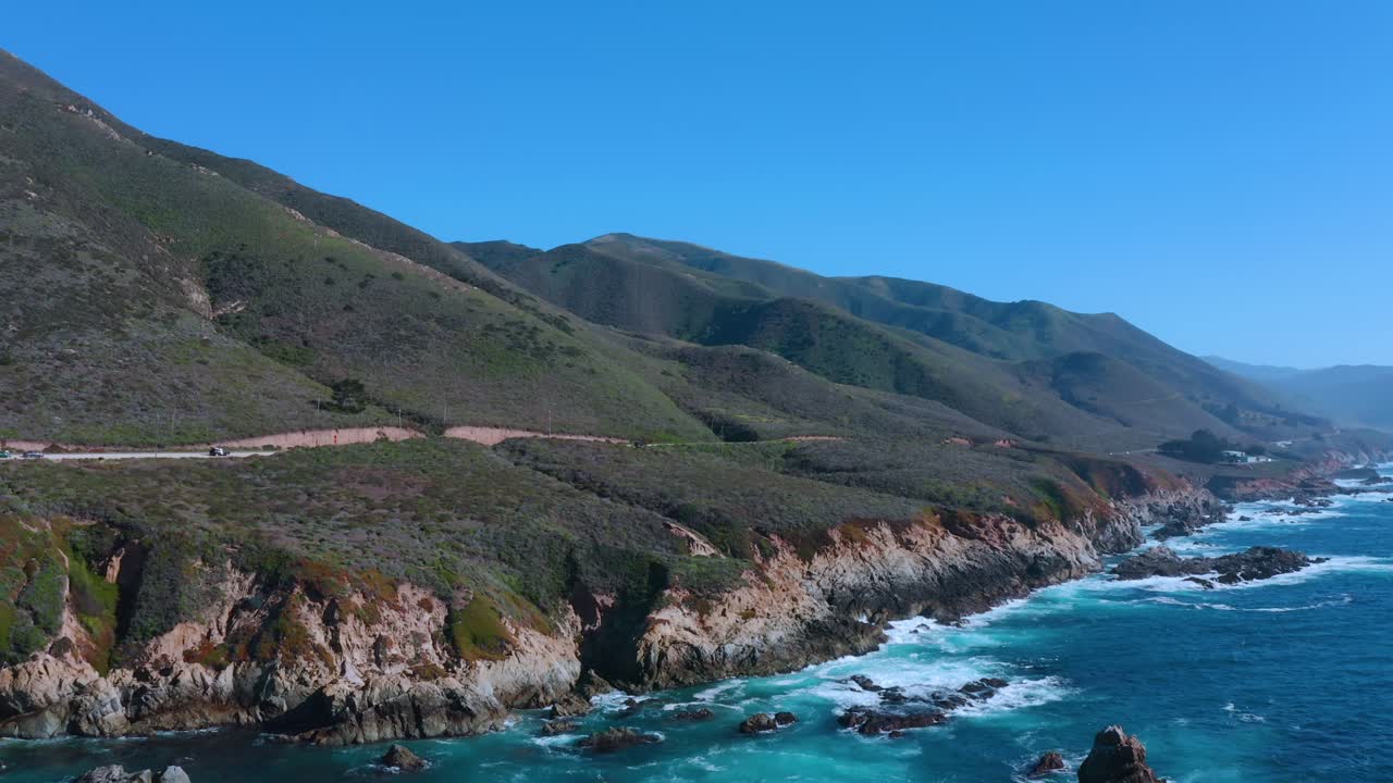 White-capped blue waves crash along the California coastline as we fly toward vehicles driving along the highway.