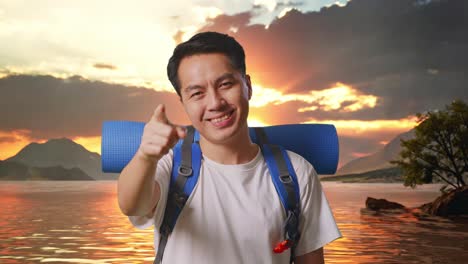 close up of asian male hiker with mountaineering backpack smiling, touching his chest, and pointing to camera at a lake