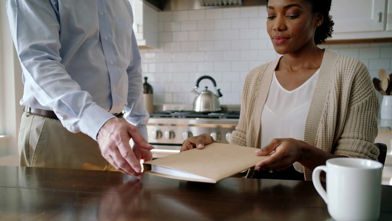 Seated woman in cardigan receiving manila folder from man coming to kitchen table with white mug