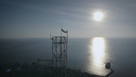 Toma-Aérea-De-Una-Torre-De-Acero-De-La-Estación-Base-Del-Transceptor-Bts-En-Una-Pequeña-Isla,-Respaldada-Por-La-Puesta-De-Sol,-Rodeada-Por-El-Océano-Azul-Por-La-Tarde