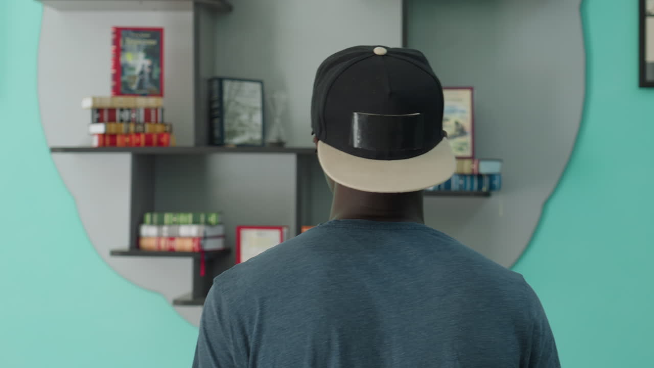 Back view of man in casual t-shirt and cap standing indoors, facing wall mounted shelves with neatly arranged books and framed art, creating calm atmosphere of quiet observation