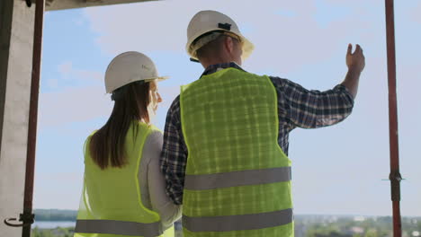 Back-turned-architect-and-technician-at-construction-site-reviewing-office-blueprints