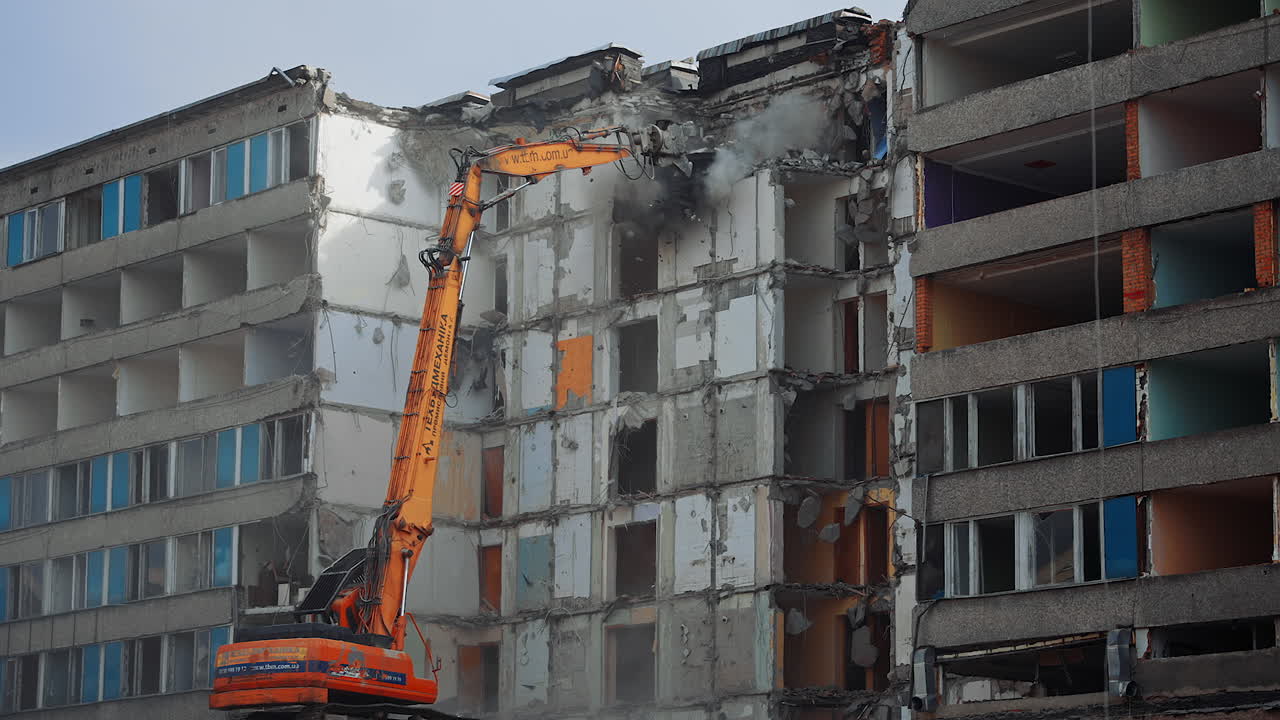 Collapse of the ruined roofs of an old house. Excavator grabber tears off the pieces of the roof. Blue sky at the backdrop.