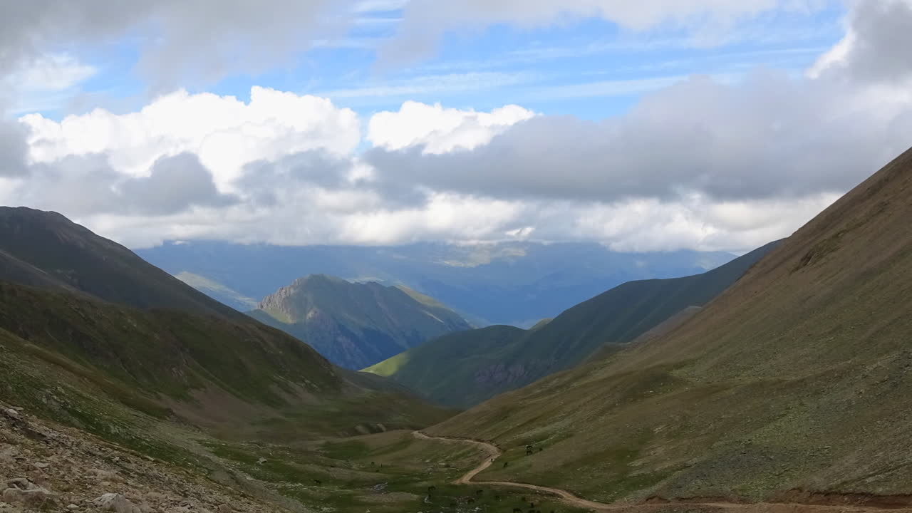 Timelapse panoramico di un'ampia valle montana con una strada sterrata tortuosa, pendii verdi e nuvole in movimento sopra le creste nel Caucaso, Russia, d'estate all'aperto