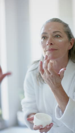 vertical video: applying face cream, mature woman in bathrobe caring for skin at home