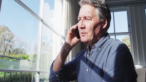 Happy-senior-caucasian-man-sitting-in-sunny-living-room-talking-on-smartphone-smartphone-and-smiling